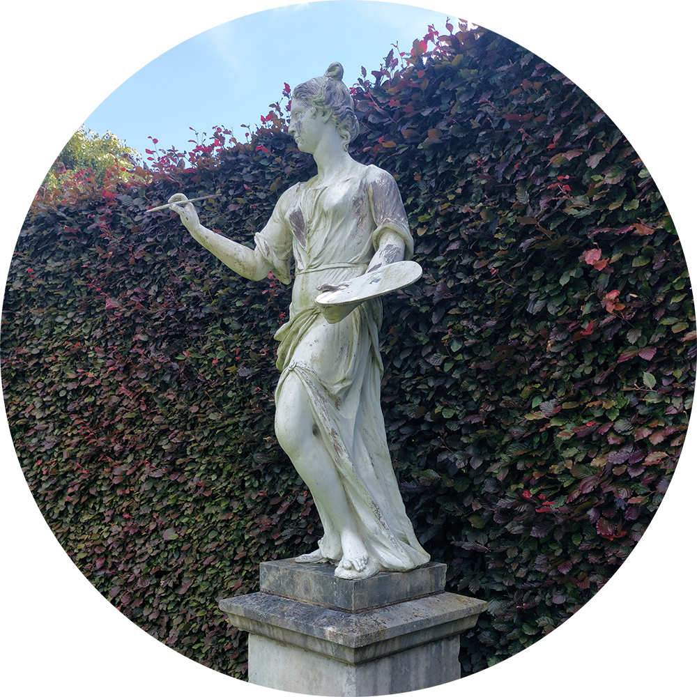 Image: A life-sized lead statue of a female figure on a plinth in front of a tall hedge of Copper beech. She is holding a paint palette in her left hand and a raised paint brush in her right. She is Apollo's muse, Painting.