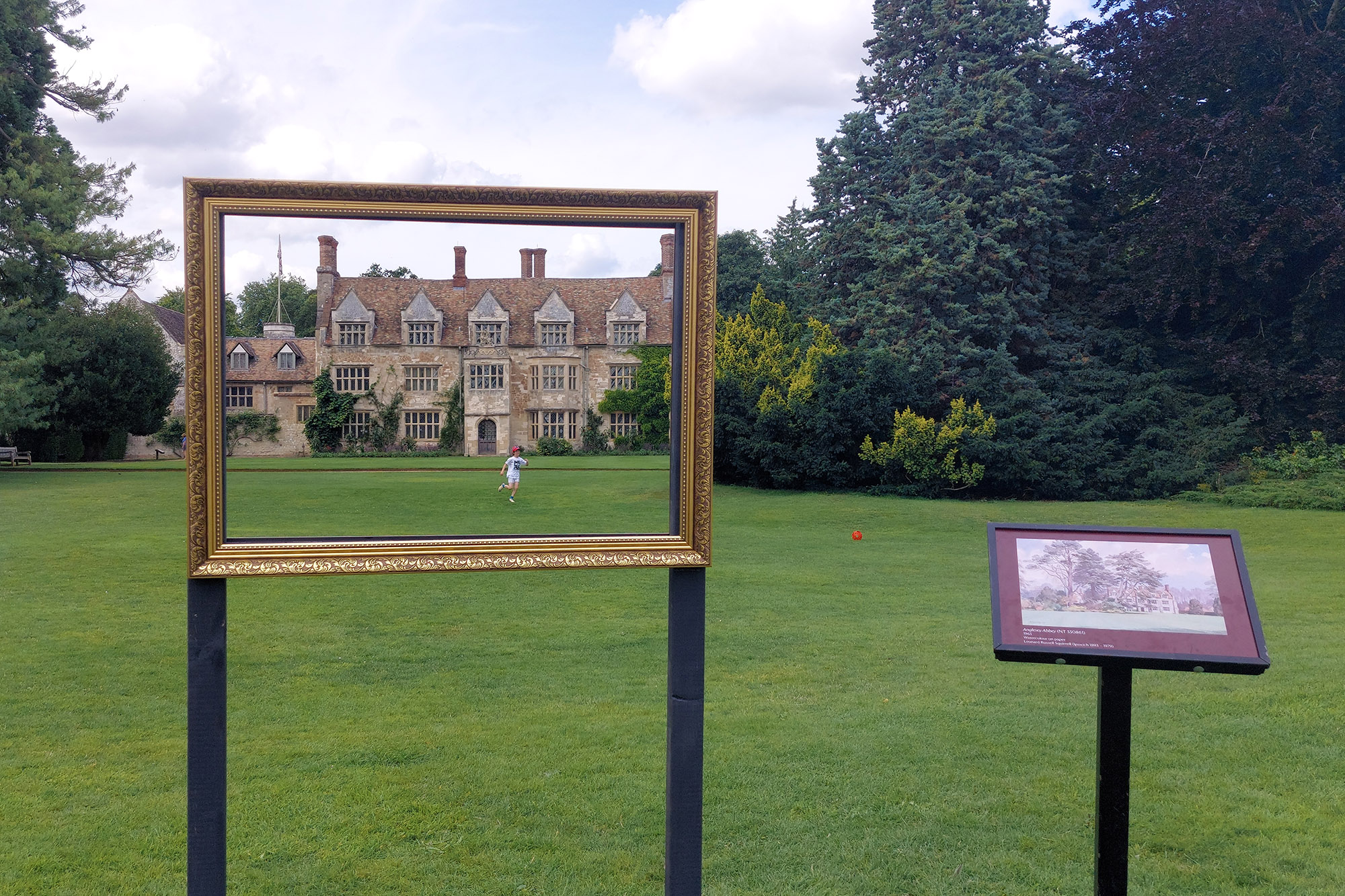 Image: An empty gilded frame captures the view of Anglesey Abbey and a child running across the lawn. Next to the frame, a board showing a painting of the scene.